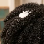 Close-up of a single tightly coiled 4C strand section with water droplets beading on the surface of the hair instead of soaking in, a dab of white conditioner sitting on top, shot in so
